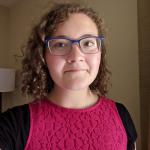A light skinned woman with shoulder length curly, brown hair. She has blue glasses and is wearing a red dress.